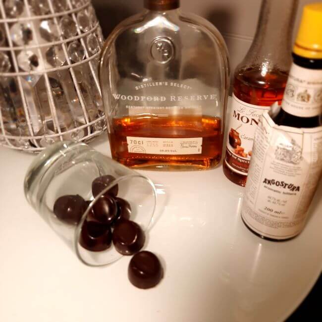 glass on side with chocolates spilling out, bottles of bourbon, bitters and syrup from Cocktail Chocolates Drawer Box